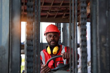Depo işçisi üniformalı ve kasklı Afro-Amerikan bir adam limandaki konteynırı kaldırmak için dizel bir forklift kullanıyor. Gülümseyen mutlu ticari taşımacılık konsepti kargo ithalat-ihracat