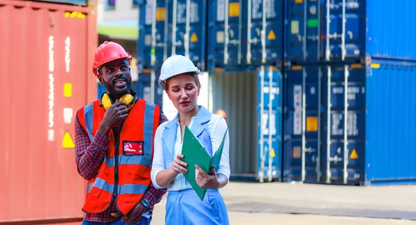 Afro-Amerikalı erkek çalışanlar veya yöneticiler ve beyaz kadınlar, konteynırları, konteynerleri, konseptleri, depoyu, ithalatı, ihracatı, lojistik sektörünü kontrol etmek için kask takın, çalışın, konuşun, belgelerinizi saklayın.