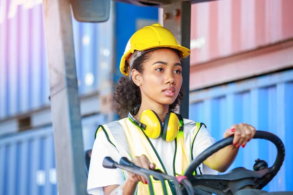 Üniformalı ve kasklı Afro-Amerikalı bir kadın limanda malları yüklemek için konteynırları kaldırmak için dizel bir forklift kullanıyor. Ticari kargo taşımacılığı konsepti, yükleme lojistiği, ithalat-ihracat.