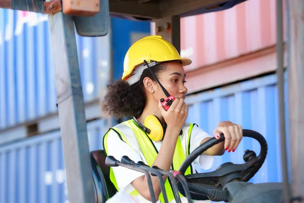 Üniformalı ve kasklı Afro-Amerikalı bir kadın limana mal taşımak için konteynır kaldırmak için forklift kullanıyor. Ticari kargo. İletişim kurmak için radyo kullanmak. Lojistik, ithalat-ihracat kavramı.