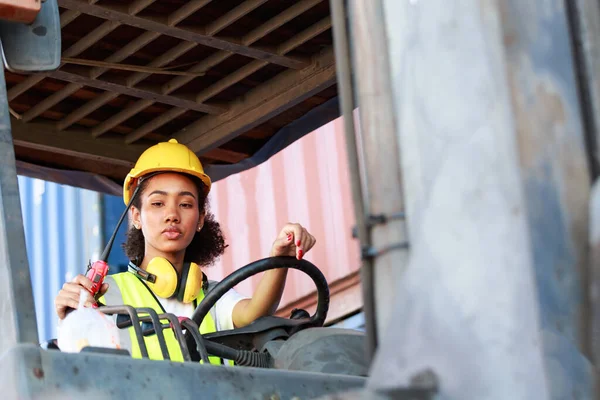 Üniformalı ve kasklı Afro-Amerikalı bir kadın limanda malları yüklemek için konteynırları kaldırmak için dizel bir forklift kullanıyor. Ticari kargo taşımacılığı konsepti, yükleme lojistiği, ithalat-ihracat.