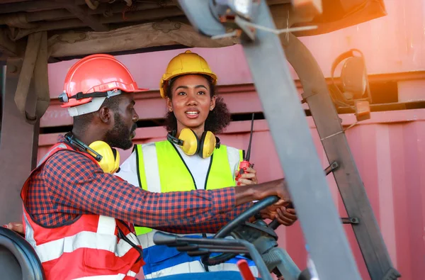 Depoda çalışan Afro-Amerikalı üniformalı kadın ve erkekler forklift kullanarak limandaki bir konteynırı kaldırıyor ve iş çıkartıyorlar. Konsept, takım, ticari nakliye. kargo ithalat-ihracatı