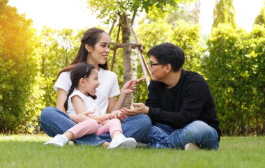 Asian family Bright and cute daughter is relaxing by running in the clear blue sky lawn and green trees, behind her parents, are taking care of them. Happy family relationship concept, lifestyle.