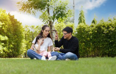 Asian family Bright and cute daughter is relaxing by running in the clear blue sky lawn and green trees, behind her parents, are taking care of them. Happy family relationship concept, lifestyle.
