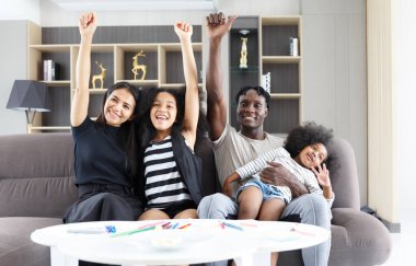 Kanepede oturan çok ırklı mutlu bir aile ve güzel bir kız çocuğu tebrik etmek için elini kaldırıyor. Çift gülümsüyor ve kameraya bakıyor. Asyalı anne ve siyah baba evde birlikte rahatlıyor.