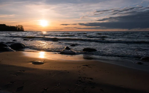 Gün batımında güneşin altında bulutlarla dolu güzel bir gökyüzünün altında, alçak köpüklü deniz dalgaları büyük taşlarla dolu vahşi kumlu bir plaja doğru koşuyor..