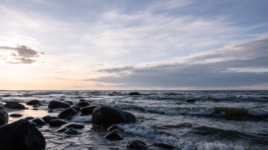 Alçak köpüklü deniz dalgaları büyük granit taşlarla dolu bir kumsalda ritmik bir şekilde ilerler. Arka ışıklandırma, akşam güneşi bulutların ve sislerin arasından doğuyor..