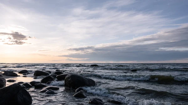 Alçak köpüklü deniz dalgaları büyük granit taşlarla dolu bir kumsalda ritmik bir şekilde ilerler. Arka ışıklandırma, akşam güneşi bulutların ve sislerin arasından doğuyor..