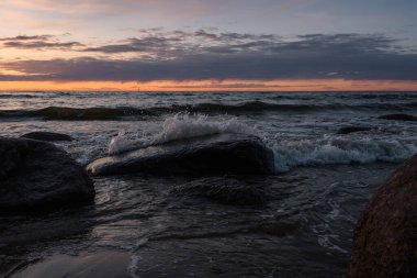 Gün batımında, karanlık ve güzel bulutlu bir gökyüzünün altında, deniz dalgaları kıyı granit kaya parçalarına ayrılır, sprey ve köpüğü yükseltir..