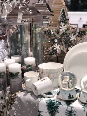 Vitrindeki bir masanın Noel süslemeleri. Beyaz ve gri renklerde. Mumlar, kupalar, sofra takımı ve kar topu.