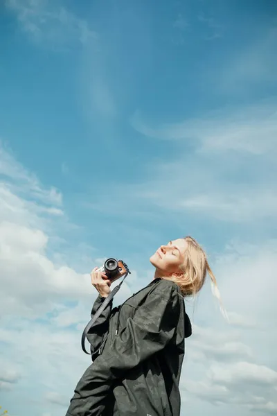 Mujer rubia joven en camisa caqui con una capucha que sostiene la cámara delante del cielo azul al aire libre — Foto de Stock