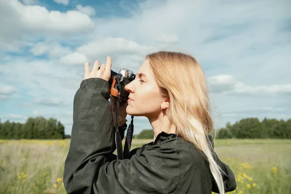 Mujer rubia joven en camisa caqui con una capucha tomando una foto con una cámara delante del cielo azul al aire libre — Foto de Stock