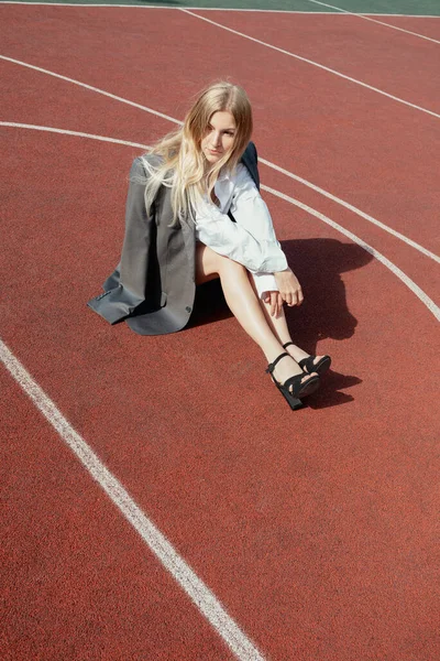Young blonde female wearing heels and jacket, sitting on a backetball sport field Royalty Free Stock Images