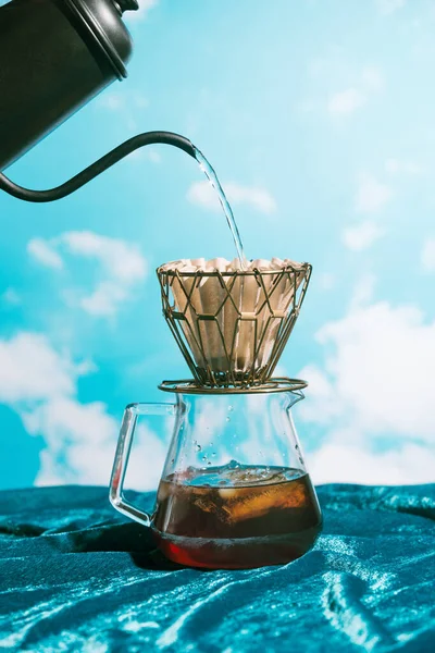 Water pouring into metall dripper with paper filter and iced coffee in front of blue sky, alternative brewing Stock Photo