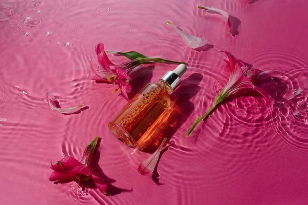 Vitamin narutal serum in a glass bottle in water surrounded by flowers on pink Stock Photo