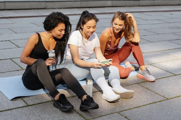 Tre donne sportive che si divertono e ridono guardando smartphone dopo l'allenamento di fitness urbano. Immagine Stock