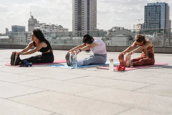 Three sporty women sitting on fitness mat and streching their legs. Stock Photo