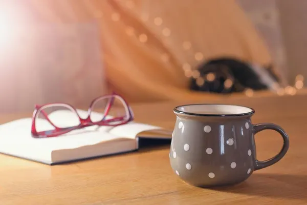 Cup of hot tea and book on the table, cozy warm home interior with sleeping cat on the background