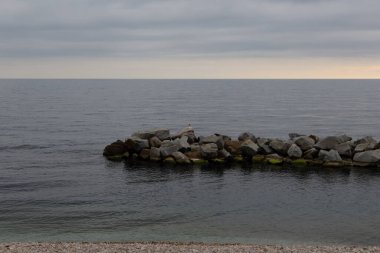 Denizde şafak söküyor, sudaki kayalardan bir tepe görünüyor ve ufukta yükselen güneşten gelen kırmızı refleks var.