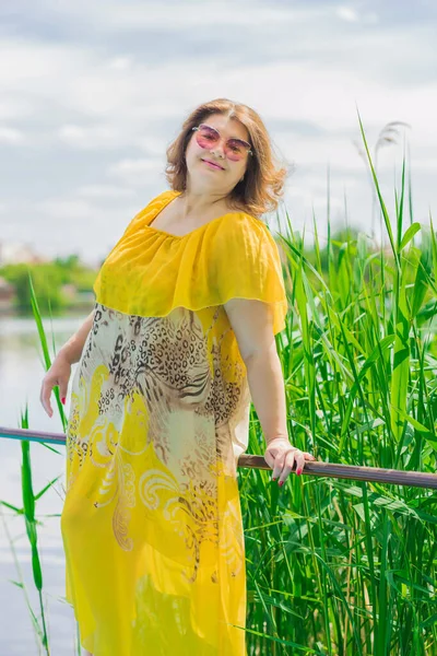 Plus size European or American woman at nature in yellow romantic dress and pink hat, enjoy the holidays. Life of people xl size, happy nice natural beauty woman. Concept of overweight