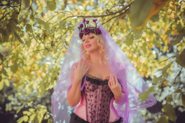 Pembe korseli Cadılar Bayramı gelini, gotik çiçekli çelenk ve doğa arka planında peçe, parti için kıyafetler.