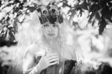 Pembe korseli Cadılar Bayramı gelini, gotik çiçekli çelenk ve doğa arka planında peçe, parti için kıyafetler.