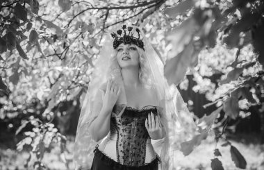 Pembe korseli Cadılar Bayramı gelini, gotik çiçekli çelenk ve doğa arka planında peçe, parti için kıyafetler.