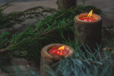 Two candles in wooden candlesticks on a wooden table between the green branches of pine and thuja. Bright fire of orange candles-tablets. Festive decor for a wedding, Valentine's Day or birthday.