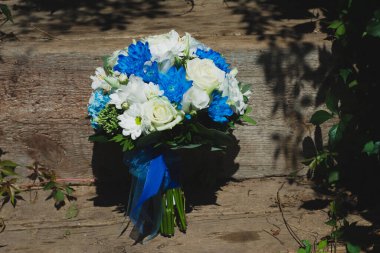 Düğün buketi papatyalar, beyaz güller, mavi kasımpatılar ve güneşli bir yaz gününde ahşap merdivenlerde mavi kurdeleye sarılı yeşil yapraklar. Mutlu bir günde gelinin düğün aksesuarı.