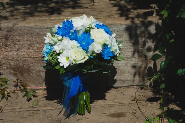 Düğün buketi papatyalar, beyaz güller, mavi kasımpatılar ve güneşli bir yaz gününde ahşap merdivenlerde mavi kurdeleye sarılı yeşil yapraklar. Mutlu bir günde gelinin düğün aksesuarı.