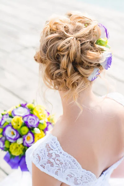Saçlarında östoma çiçekleri olan sarışın gelin güneşli bir günde elinde mor ve sarı çiçeklerden bir buket tutuyor. Bir düğün için saç stili.