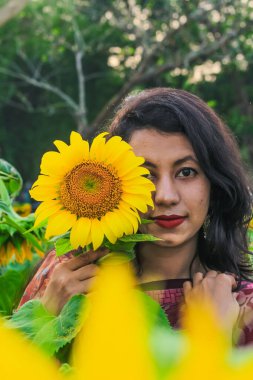 Güzel genç kız ayçiçeği tarlasında doğanın tadını çıkarıyor. Ayçiçeği tarlasında bir kız. Parkta gülümseyen ve yüzünü ayçiçeğiyle kaplayan bir kız..