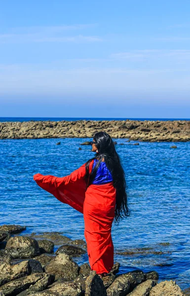 Young women wearing a red saree on the beach. Girl in traditional Indian sari among the rocks and enjoying the freedom and the sunset or sunrise. Beautiful Indian woman on the beach wearing a sari.