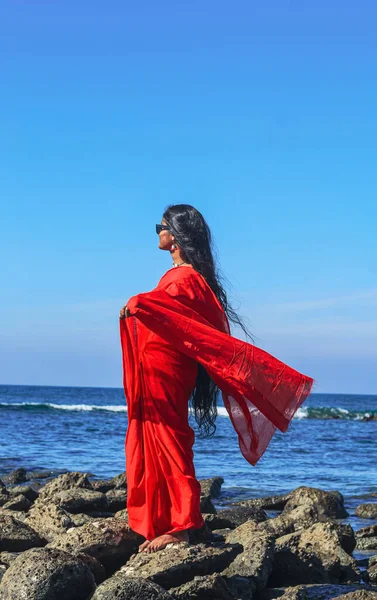 Young women wearing a red saree on the beach. Girl in traditional Indian sari among the rocks and enjoying the freedom and the sunset or sunrise. Beautiful Indian woman on the beach wearing a sari.