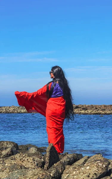 Young women wearing a red saree on the beach. Girl in traditional Indian sari among the rocks and enjoying the freedom and the sunset or sunrise. Beautiful Indian woman on the beach wearing a sari.