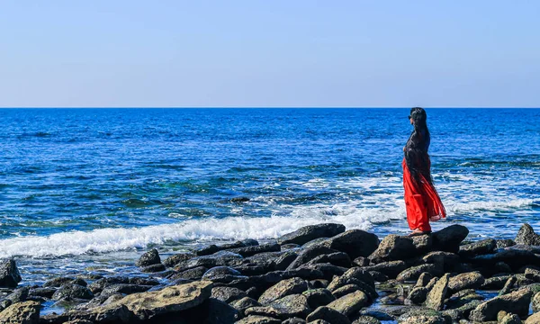 Young women wearing a red saree on the beach. Girl in traditional Indian sari among the rocks and enjoying the freedom and the sunset or sunrise. Beautiful Indian woman on the beach wearing a sari.