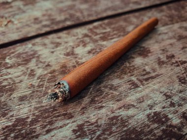 Detail of a lit marijuana or weed joint pictured on a brown wooden table outdoors.