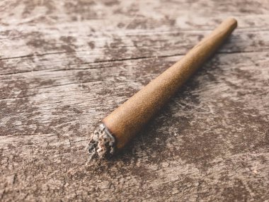 Detail of a lit marijuana or weed joint pictured on a brown wooden table outdoors.