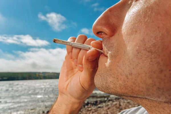Detail of a man smoking a marijuana or weed joint relaxing on the shore of a lake.