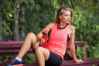 Atletik yapılı bir kız spor antrenmanından sonra su içer ve parktaki bir bankta dinlenir..