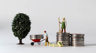 Business concept with coins and miniature people. A miniature man carrying coins in a cart and a miniature man standing on a pile of coins.