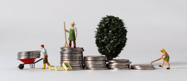 Business concept with coins and miniature people. A miniature man carrying coins in a cart and a miniature man standing on a pile of coins.