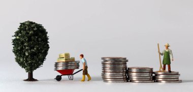 Business concept with coins and miniature people. A miniature man carrying coins in a cart and a miniature man standing on a pile of coins.