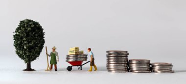 Business concept with coins and miniature people. A miniature man carrying coins in a cart and a miniature man standing on a pile of coins.