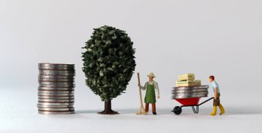 Business concept with coins and miniature people. A miniature man carrying coins in a cart and a miniature man standing on a pile of coins.