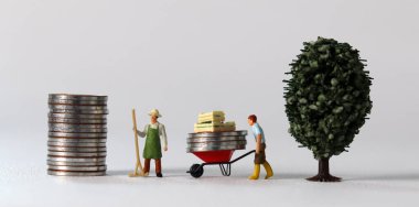 Business concept with coins and miniature people. A miniature man carrying coins in a cart and a miniature man standing on a pile of coins.