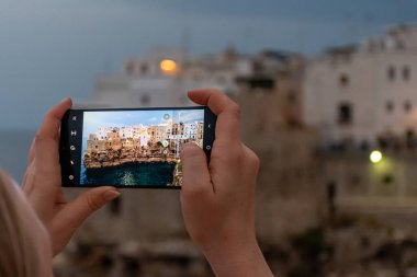 Karabasan Polignano. Kayalıklardaki kasaba, Puglia bölgesi, İtalya, Avrupa. Kayalıklardaki kasaba. Eski geleneksel evleri, dramatik gökyüzünü, Akdeniz 'i ve sarışın turist kızın akşamları akıllı telefonuyla fotoğraf çektiği seyahat konsepti.