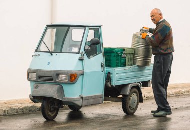 Cisternino, İtalya - 8 Kasım 2021: Puglia, İtalya 'da yeni yapılmış bir zeytinyağı fıçısı taşıyan eski model hafif maymun arabalı yaşlı çiftçi