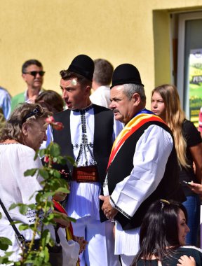 Geleneksel siyah motiflerle dikilmiş ulusal çoban elbisesi giymiş, Romanya 'da bir belediye başkanı, yuvarlak siyah bir şapka, geniş bir deri kimir ve belediye başkanı için üç renkli bir eşarp giyiyor.