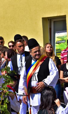 Geleneksel siyah motiflerle dikilmiş ulusal çoban elbisesi giymiş, Romanya 'da bir belediye başkanı, yuvarlak siyah bir şapka, geniş bir deri kimir ve belediye başkanı için üç renkli bir eşarp giyiyor.